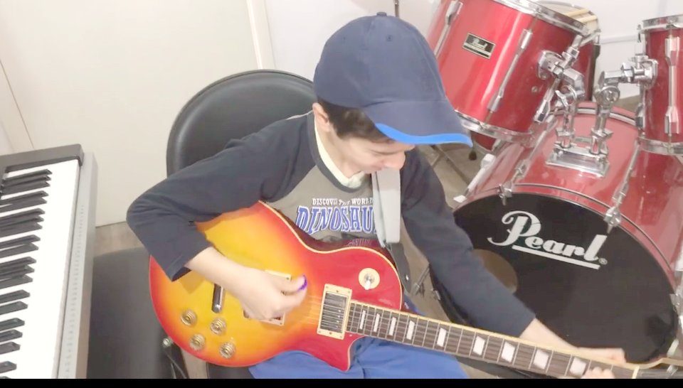 Criança tocando guitarra no estúdio de Denis Delphin, com bateria e teclado ao fundo
